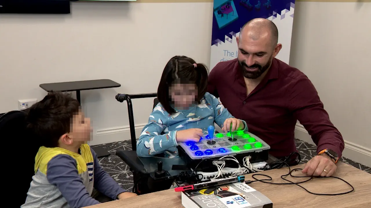 Une jeune fille utilise une manette adaptée aux besoins spécifiques, accompagnée par un adulte et un autre enfant. Cette manette fait partie d’un dispositif inclusif facilitant l’accès au jeu vidéo pour les personnes en situation de handicap.